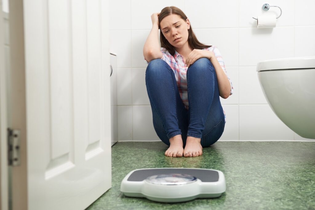 sad woman sitting on bathroom floor behind scale