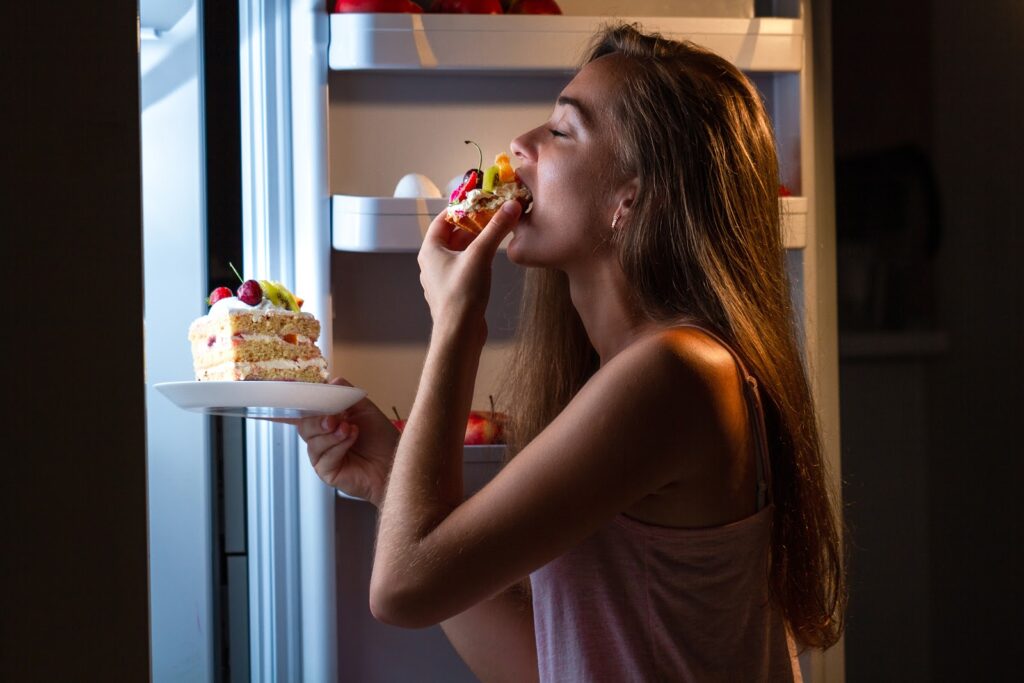 girl standing in front of an open refrigerator eating cake with her hand